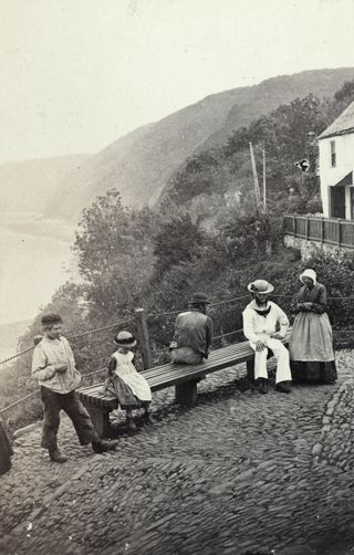 Black and white image of people in Edwardian dress sitting on a bench