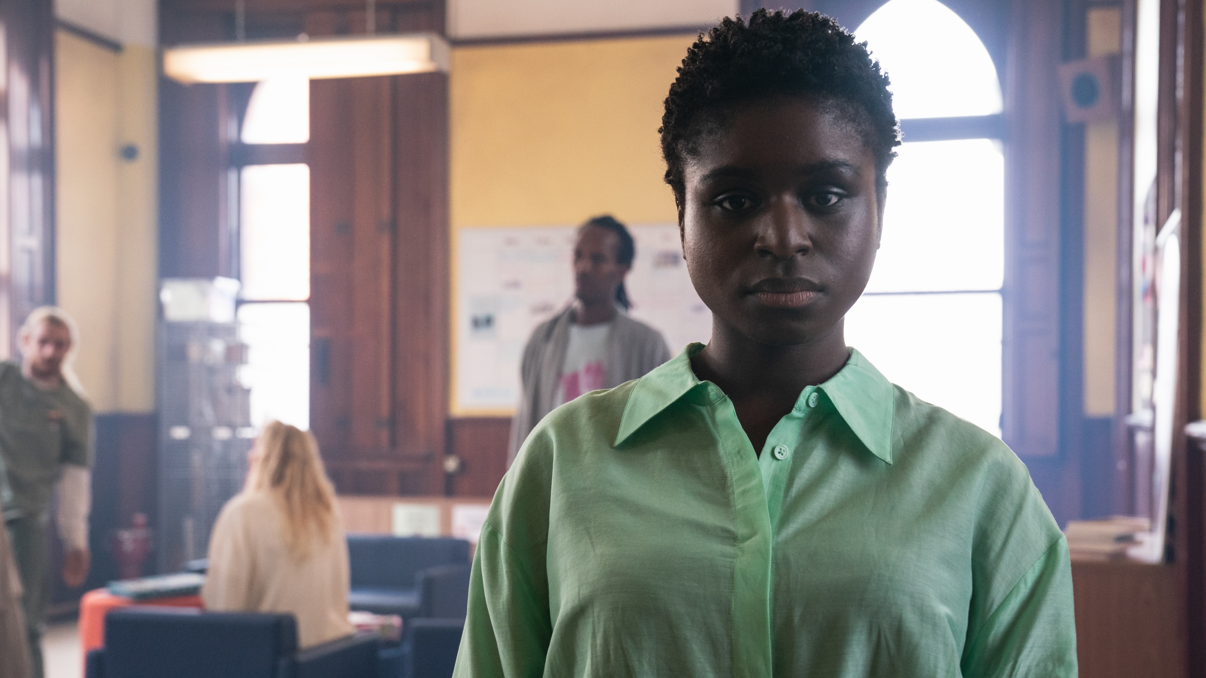 Pictured: JESSICA LAYDE as Bernadette in a green top and looking serious as patients stand behind her.