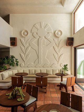 A hotel lobby punctuated by organic mid-century modern furniture in wood and softly colored vintage seating features a relief mural depicting the silhouette of a woman holding a sun-shaped talisman in between two flowers.