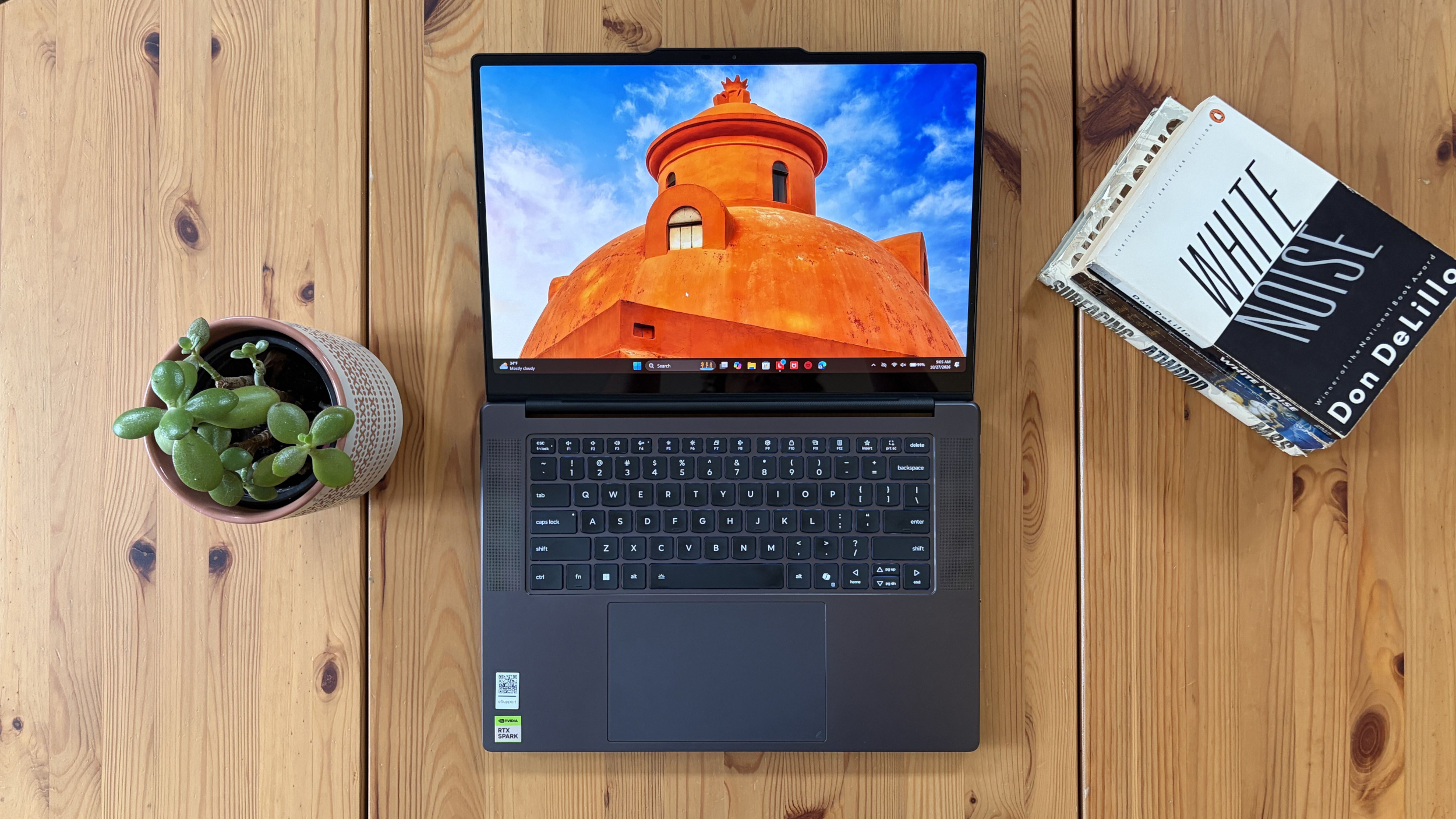 Overhead view of a Lenovo Pro 9n laptop showing a bright orange domed building, a small green succulent, and a copy of &amp;ldquo;White Noise&amp;rdquo; on a wooden desk; calm, inviting workspace.