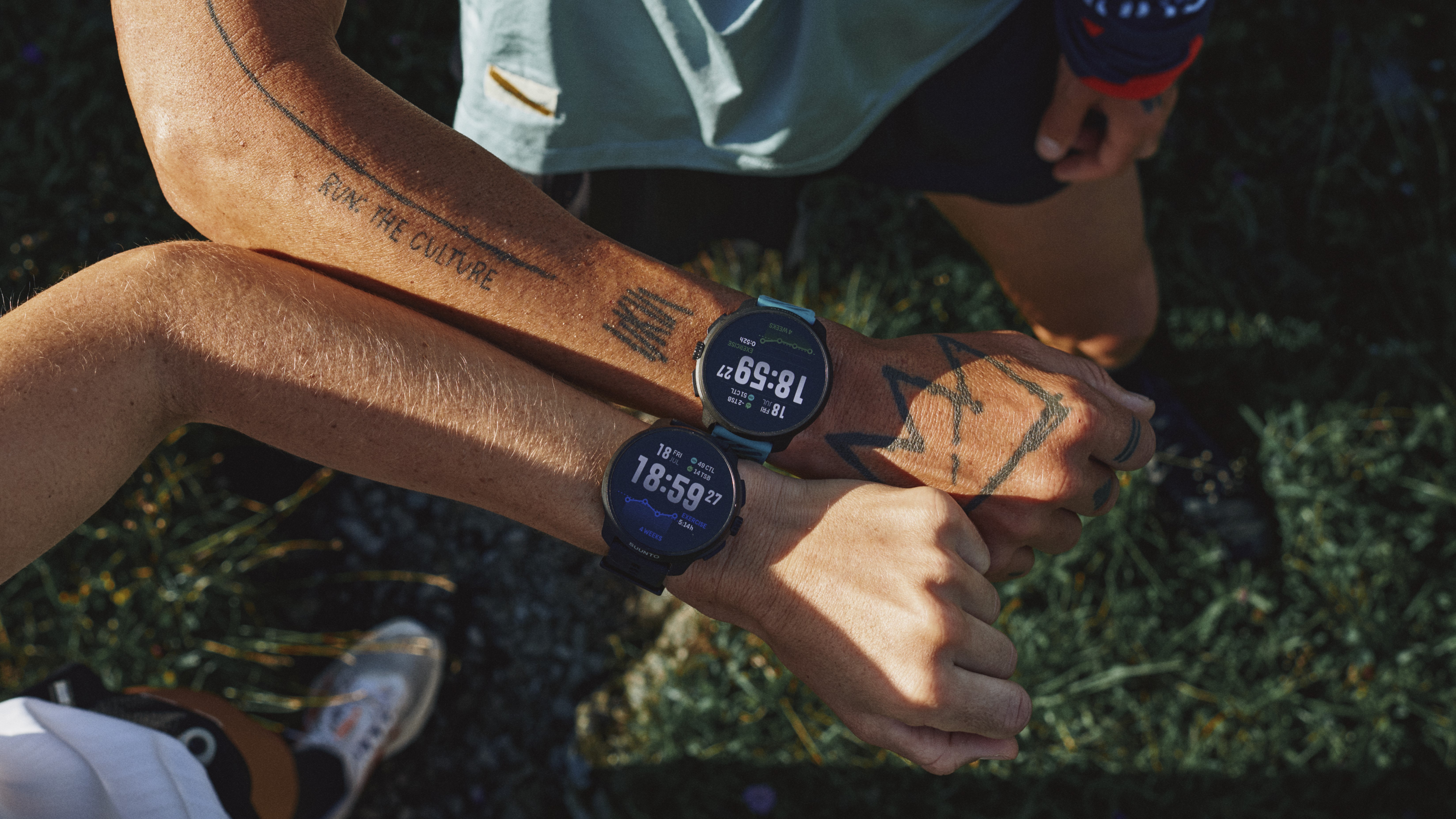 A photo from above of two runners holding their arms parallel to each other, both wearing different colors of the Suunto Race 2 running watch.