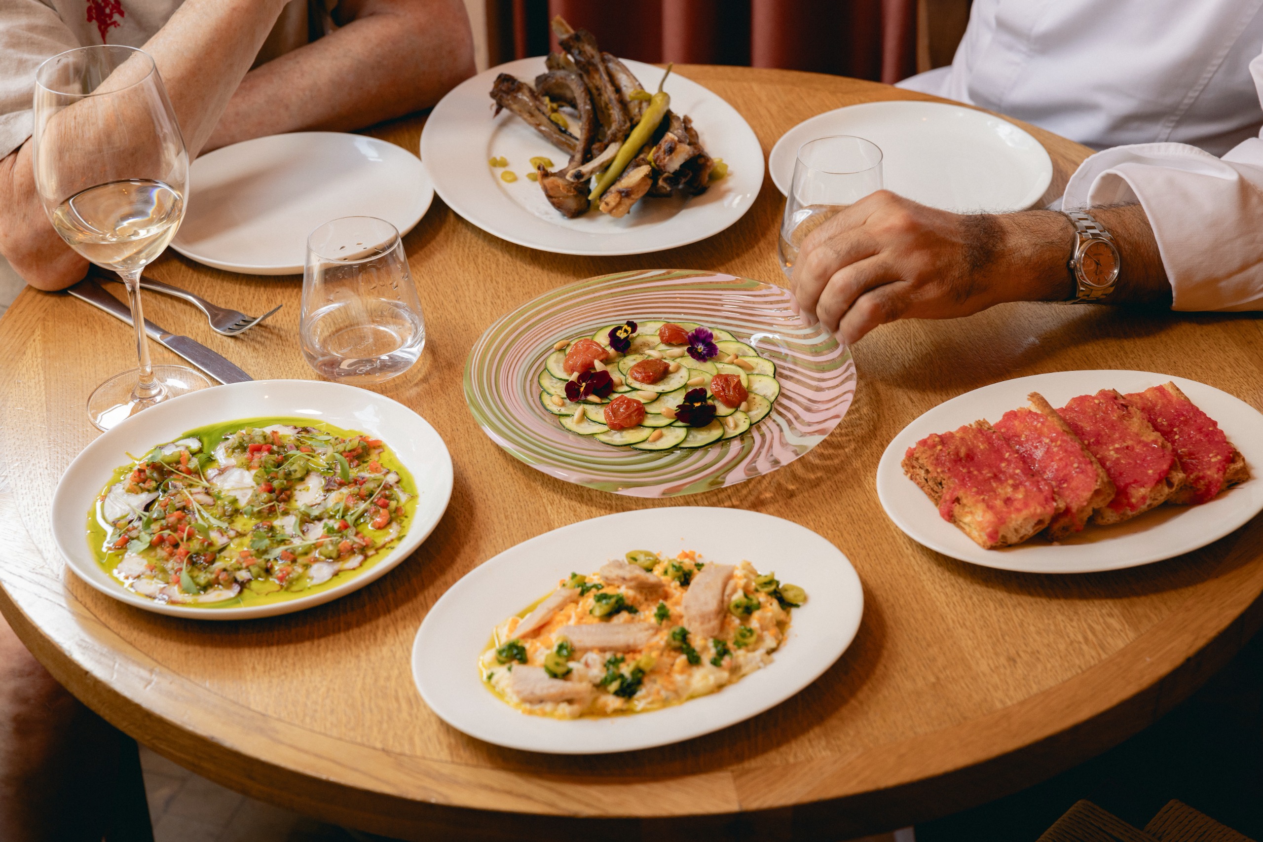 A wooden table on top of which sit ceramic and mouth-blown glass plates with pink and green wavy textures, filled with colorful food including crudo, courgette and flowers, and slices of tomato bread.