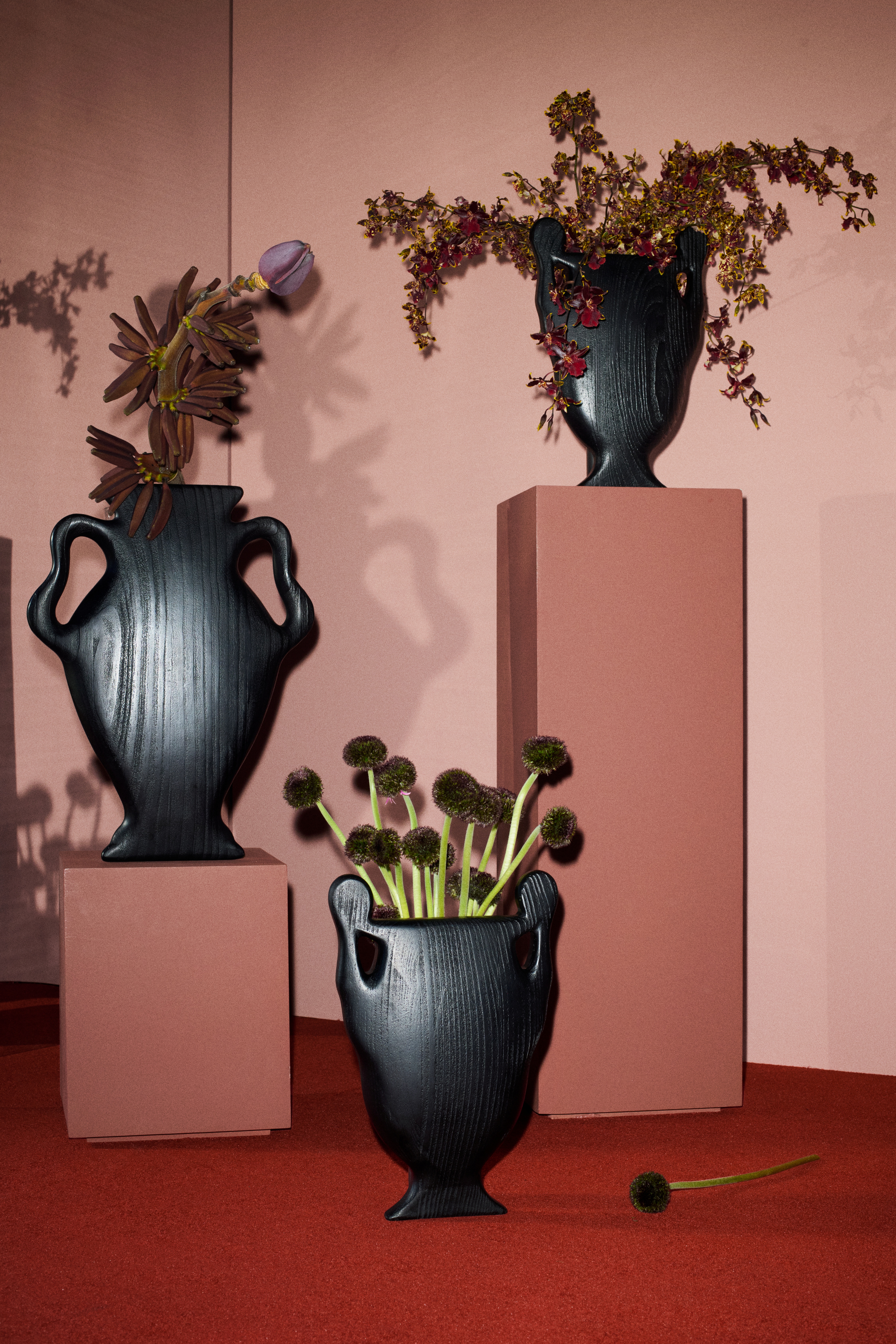 Installation shots of sculpturally designed pieces, including vases, coffee tables, armchairs, and black-and-white clothing hung up on a bordeaux rail in rooms with red or ochre carpet.