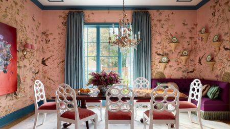 Vibrant dining room with pink bird wallpaper, teal trim, white open-back chairs around a wooden table, a purple corner banquette, and a gold chandelier.