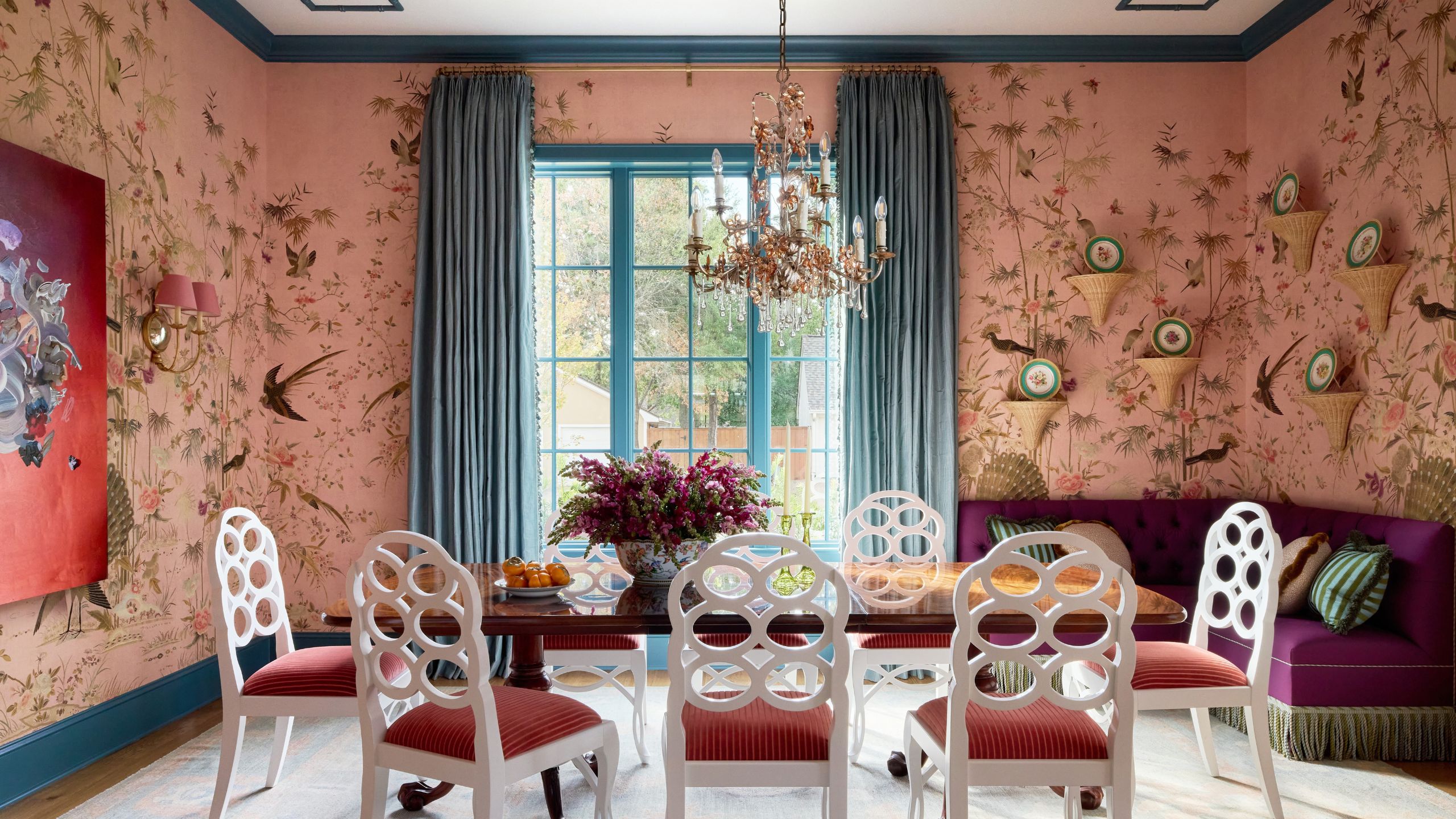 Vibrant dining room with pink bird wallpaper, teal trim, white open-back chairs around a wooden table, a purple corner banquette, and a gold chandelier.