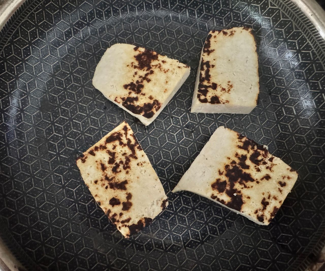 Four large chunks of tofu in a black lined frying pan, with char marks.