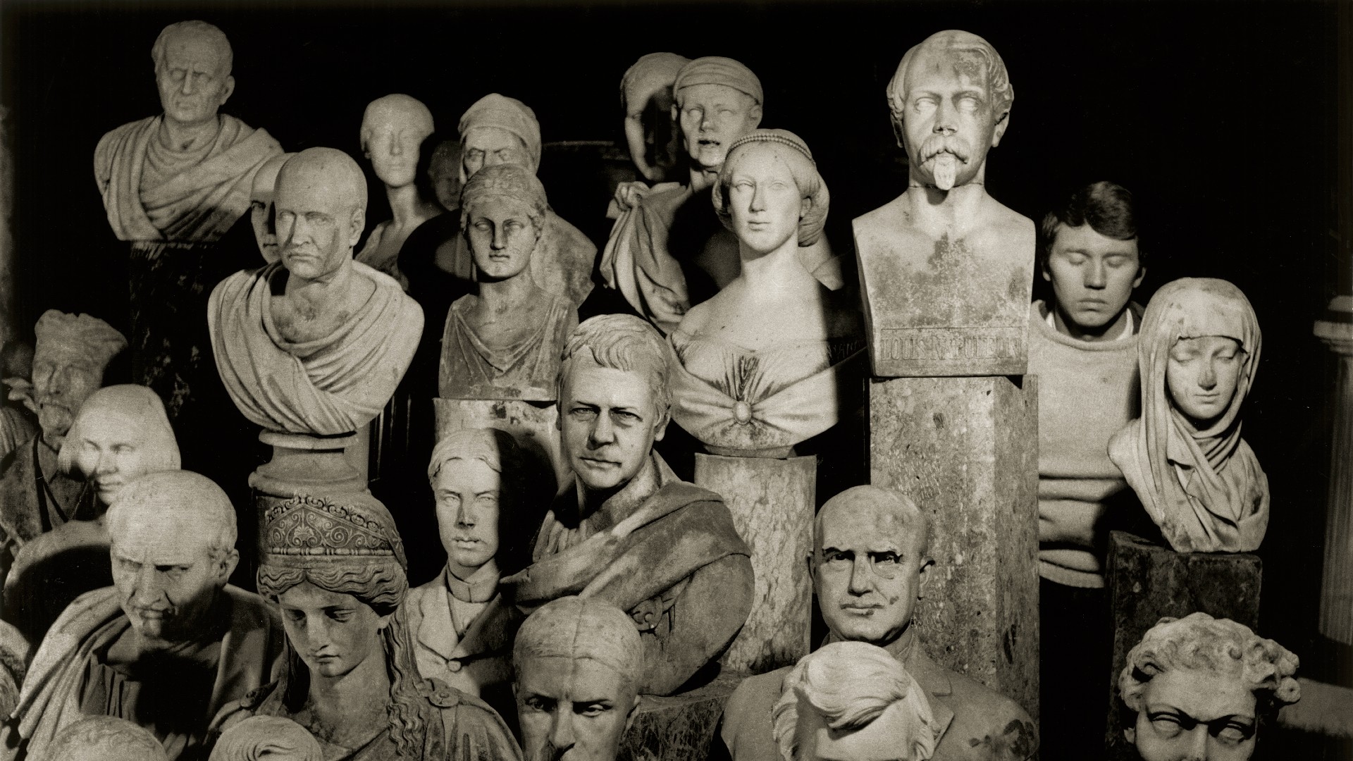 Don McCullin&rsquo;s face is partially hidden as he blends into a dense, atmospheric arrangement of historical stone busts and sculptures.