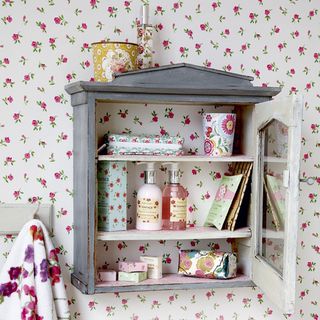 bathroom with white and pink floral wallpaper, and a grey vintage backless cabinet containing bathroom products