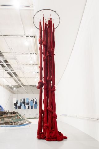 A monumental, fabric installation in bright red features knots and hangs from the ceiling of a white gallery.