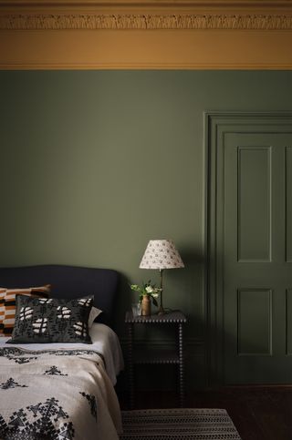 green bedroom with green walls and door, ochre picture rail and ceiling, black bed dark grey nightstand, rug, patterned lamp, patterned blanket and pillows