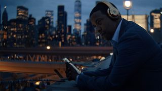 Denzel Washington in a still from Highest 2 Lowest as he leans against a wall wearing headphones and holding a phone against a city skyline