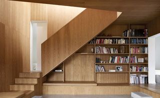 The living room is done in soft oiled oak. To the left, we see the stairs that lead to the upper floor. To the far wall, there is a built-in bookshelf filled with books.