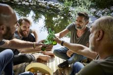 Four middle-aged male friends sit on a dock and toast with bottles of beer. They sit next to a small pond.