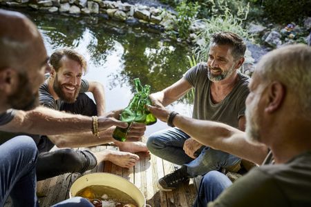 Four middle-aged male friends sit on a dock and toast with bottles of beer. They sit next to a small pond.