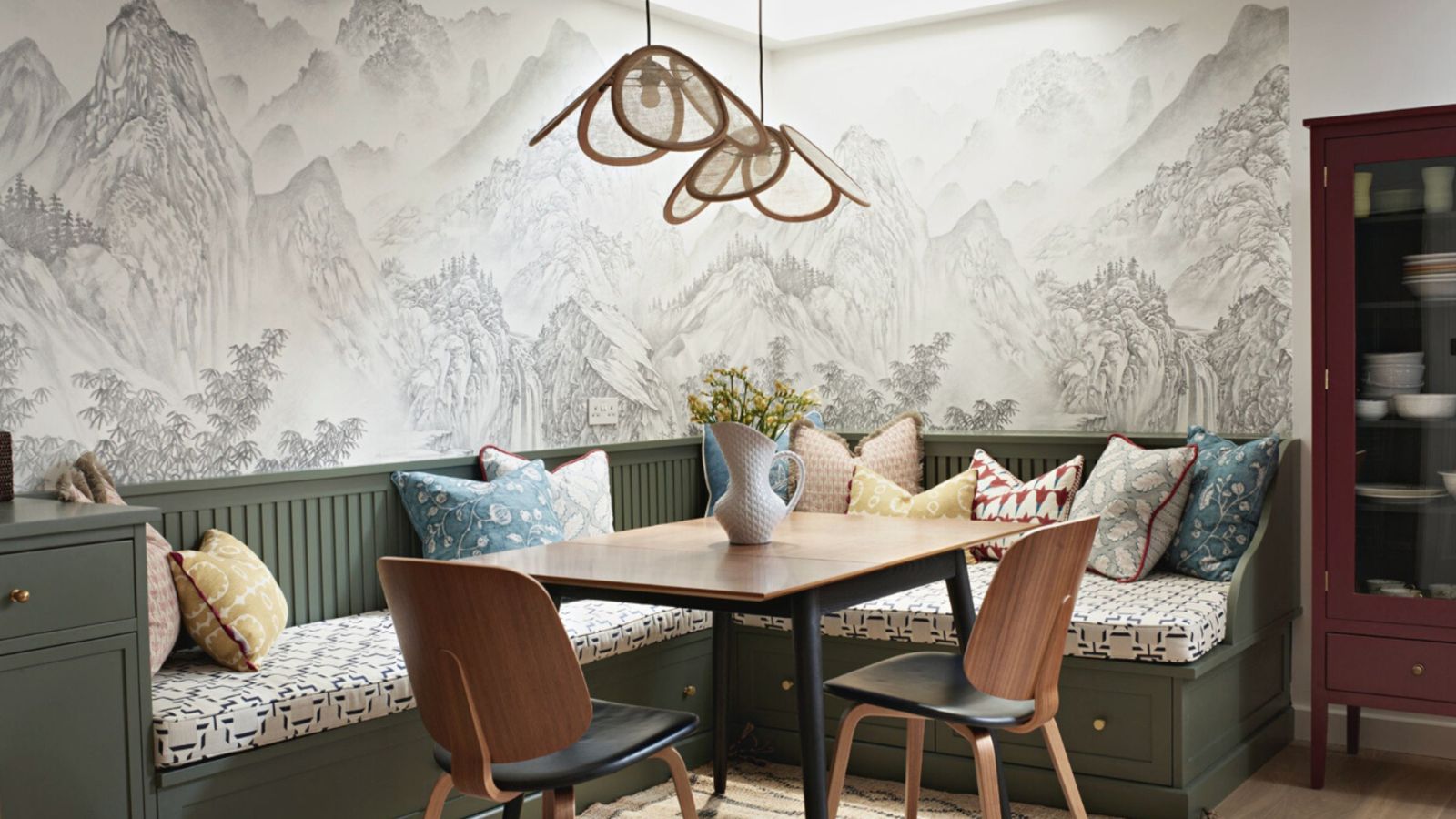 A green banquette bench behind a wooden rectangular dining table. two wooden chairs either side. Wicker lampshades hang from pendant lights above. 
