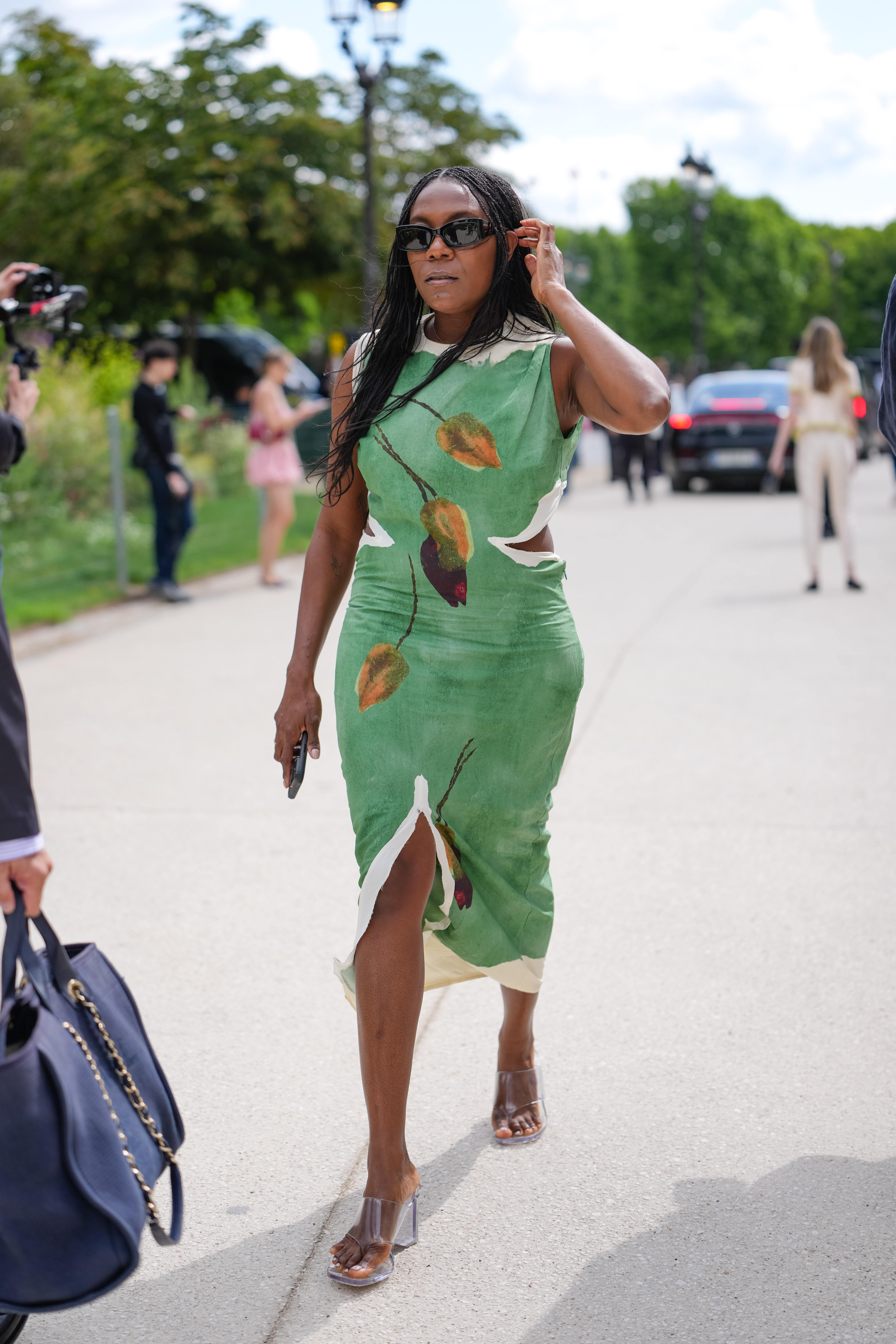 Una invitada luce un vestido sin mangas con una vibrante base verde y un estampado botánico en tonos naranja, amarillo y burdeos. El vestido tiene una silueta ajustada con escote redondo y sutiles aberturas laterales. Lleva gafas de sol oscuras y el cabello liso, cayendo sobre los hombros. El calzado consiste en sandalias abiertas en tono marrón. Una pequeña prenda negra se sostiene en una mano, a la entrada de Chanel, durante la Semana de la Moda de París - Alta Costura Otoño/Invierno 2025/2026.