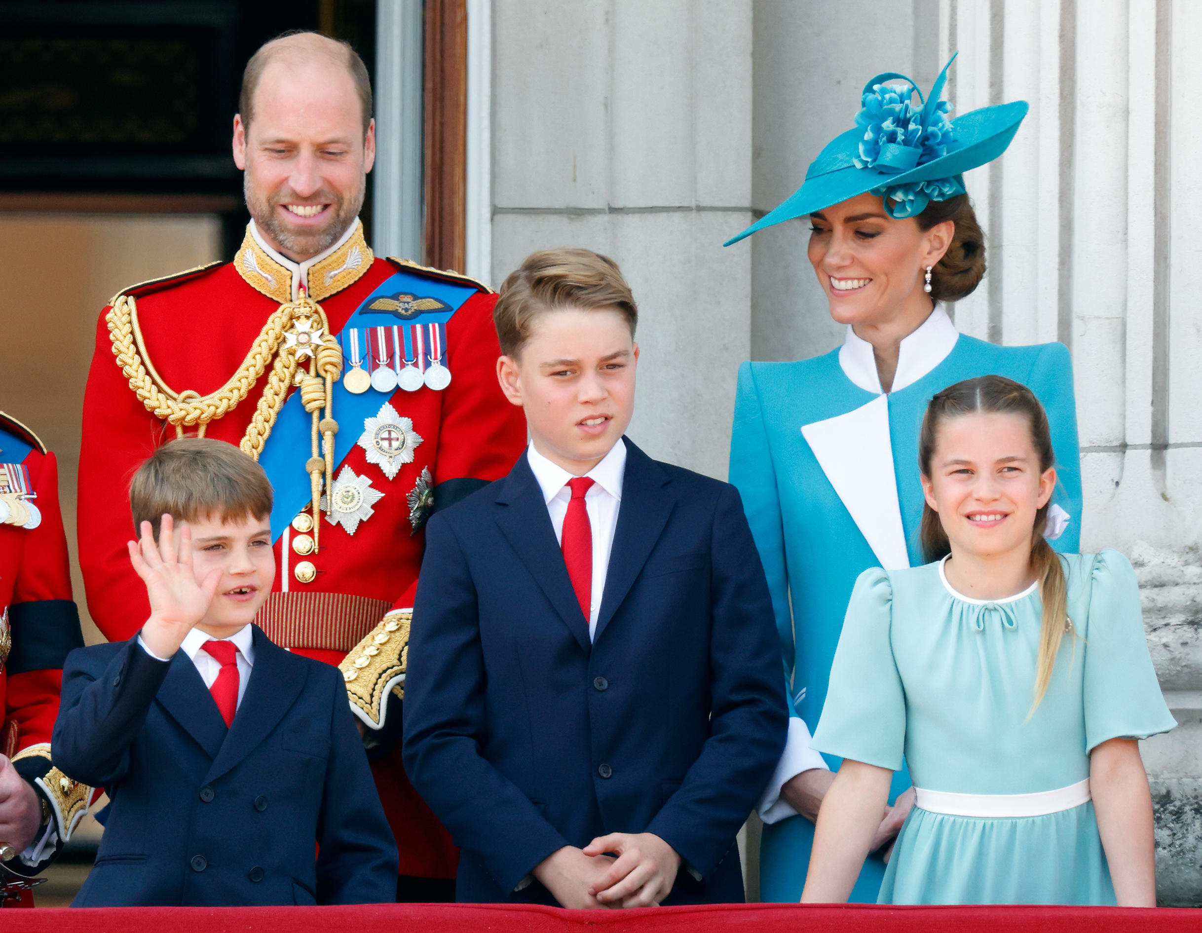 Prince William and Princess Kate smiling at Prince Louis, who is waving on the balcony next to Prince George and Charlotte