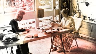 two older women sitting at a cluttered table doing arts and crafts together with a red and yellow filter