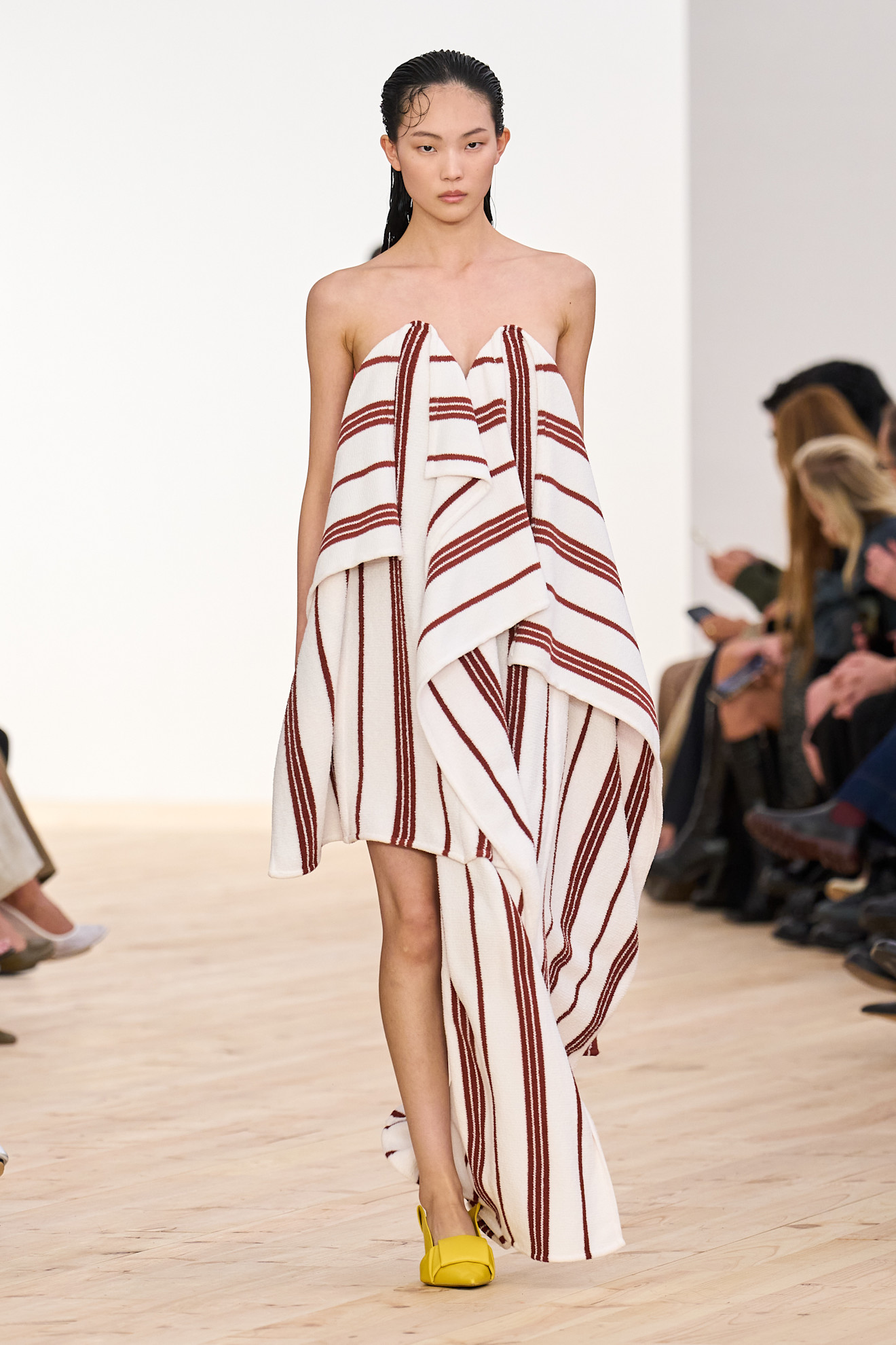 A model walking in the SS26 Loewe show is wearing a striped red-and-cream strapless dress with yellow shoes.