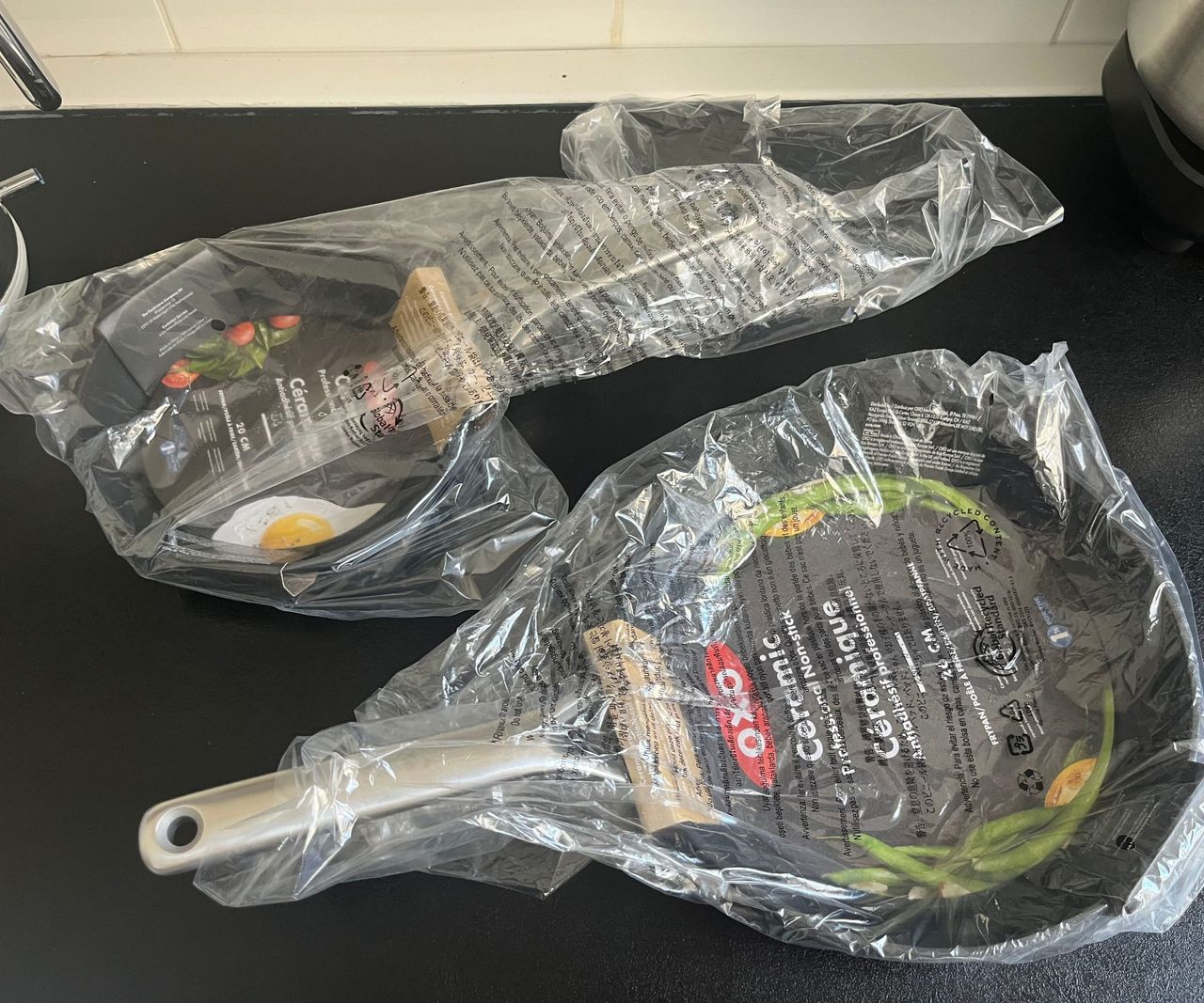 Two black ceramic frying pans on a black countertop in plastic bags.