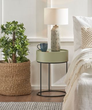 A white bedroom with a rattan floor plant pot, a circular sage green nightstand with a tall petal lamp, and a white bed with layered cream texturing