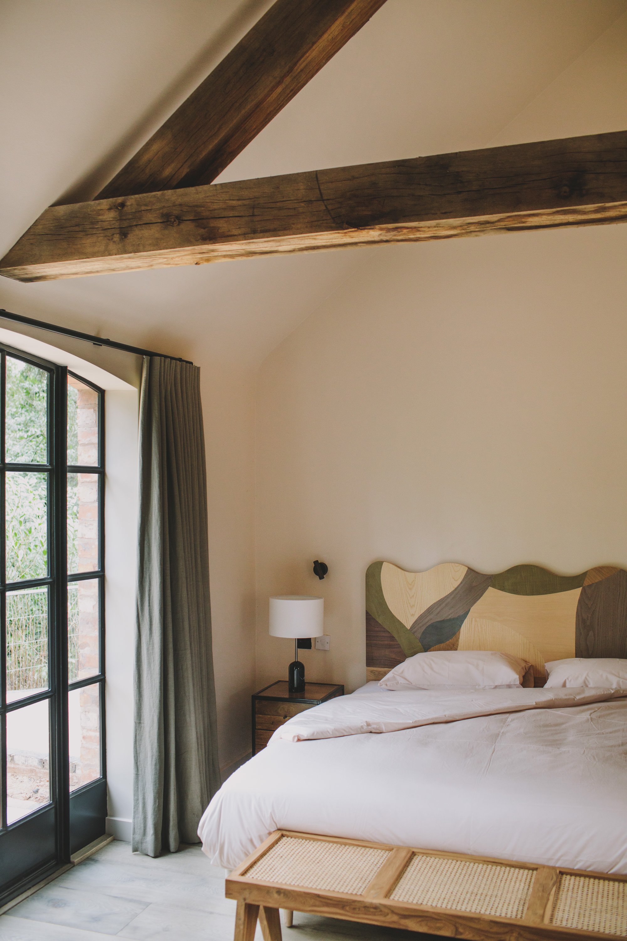 A minimally furnished, cozy hotel room features wood accents, including a pastel-shaded headboard with an abstract motif and rattan bed bench, marble floors, and a muted green and nude palette.
