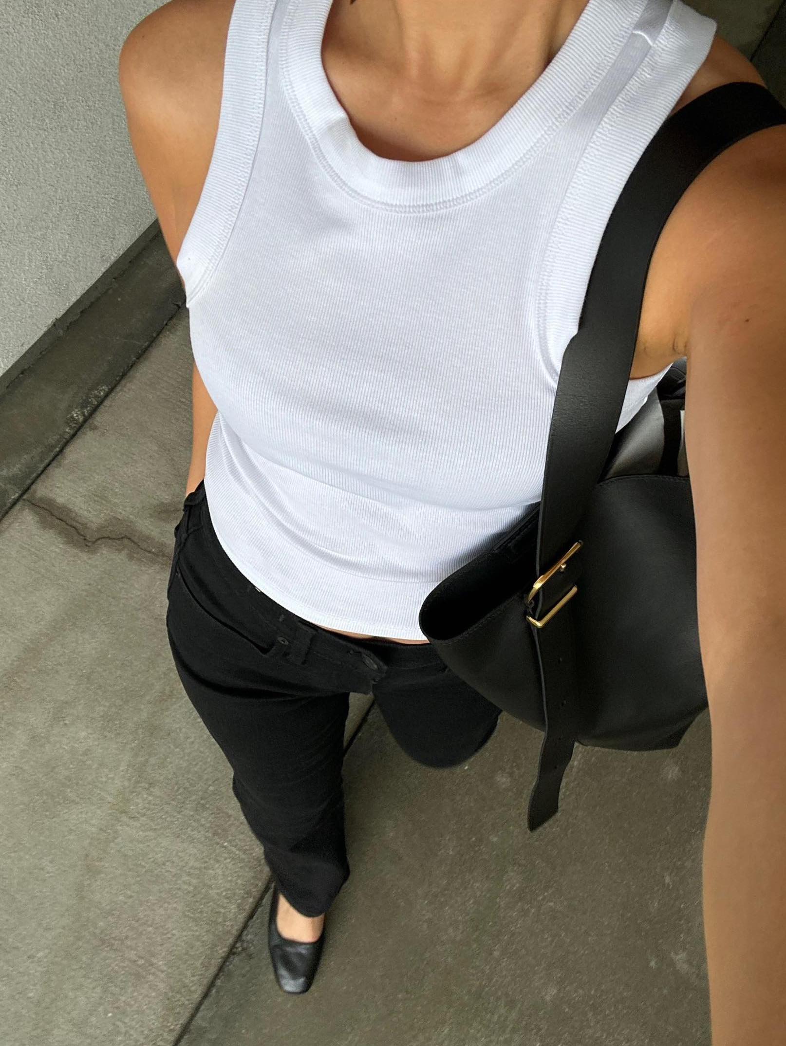 influencer Jordan Santos takes a selfie of her outfit from above wearing a white ribbed tank top, black leather tote bag, black pants, and black ballet flats