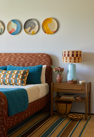 A bright and patterned bedroom with blue and ochre yellow/orange being the main color theme. The bed is a burnt orange with bedding that is beige, dark teal, and orange. There is a decorative, geometric throw pillow. There are plates hung on the wall and a wooden beside table beside the bed.