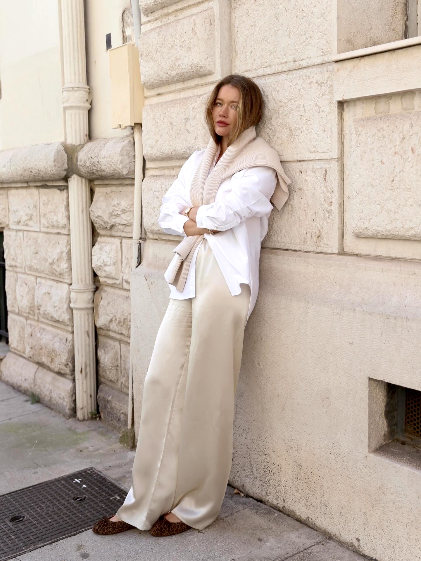style influencer Lovisa Barkman poses on a sidewalk in C&amp;ocirc;te d'Azur French Riviera wearing a white button-down shirt with a beige sweater around her shoulder, ivory satin pants, and leopard-print ballet flats