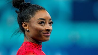 Simone Biles smiling with her hair tied into a bun, wearing a sparkly red top, standing in front of a blue background