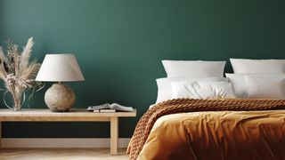 A bedroom with a green wall, a bed with an orange velvet throw and a coffee table with a lamp and vase on