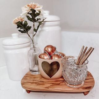 Cute coffee tray detail with heart mug filled with coffee capsules, sugar sticks in glass vessel, and flower sprig in glass bud vase on small wooden tray