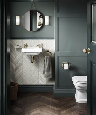 Elegant small bathroom with deep green wall panelling, a white wall-mounted sink, and brushed brass fixtures. Chevron tile wainscoting, herringbone wood flooring.