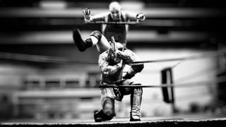 Black and white photograph of Billy Moshpit dropping a leg lariat across a kneeling luchador, draped across the middle rope, at a UWI professional wrestling event