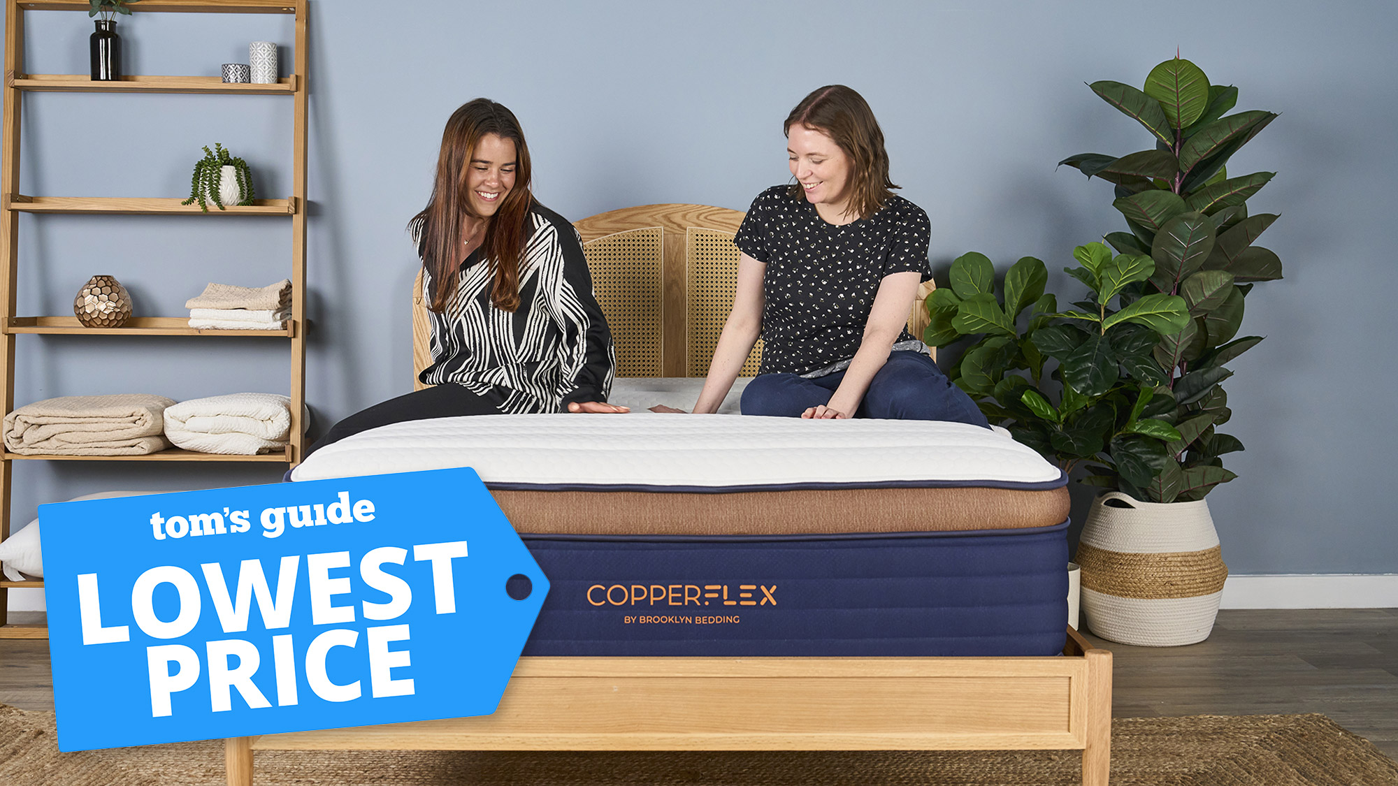 Two women sitting on the Brooklyn Bedding CopperFlex Pro Hybrid mattress in a bedroom, with a Tom's Guide label overlaid saying 'LOWEST PRICE'