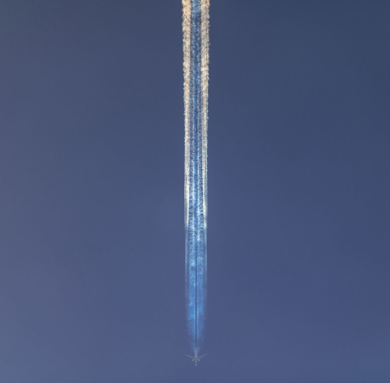rainbow contrails behind an airplane in front of a blue sky.