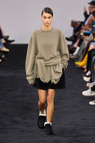 JW Anderson fall/winter 2024 model wears a beige sweater tied around another beige sweater with a black skirt and black shoes.