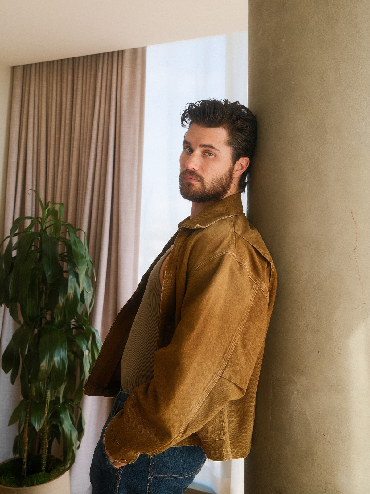 Chase Stokes leans against a wall wearing a brown chore jacket and jeans.