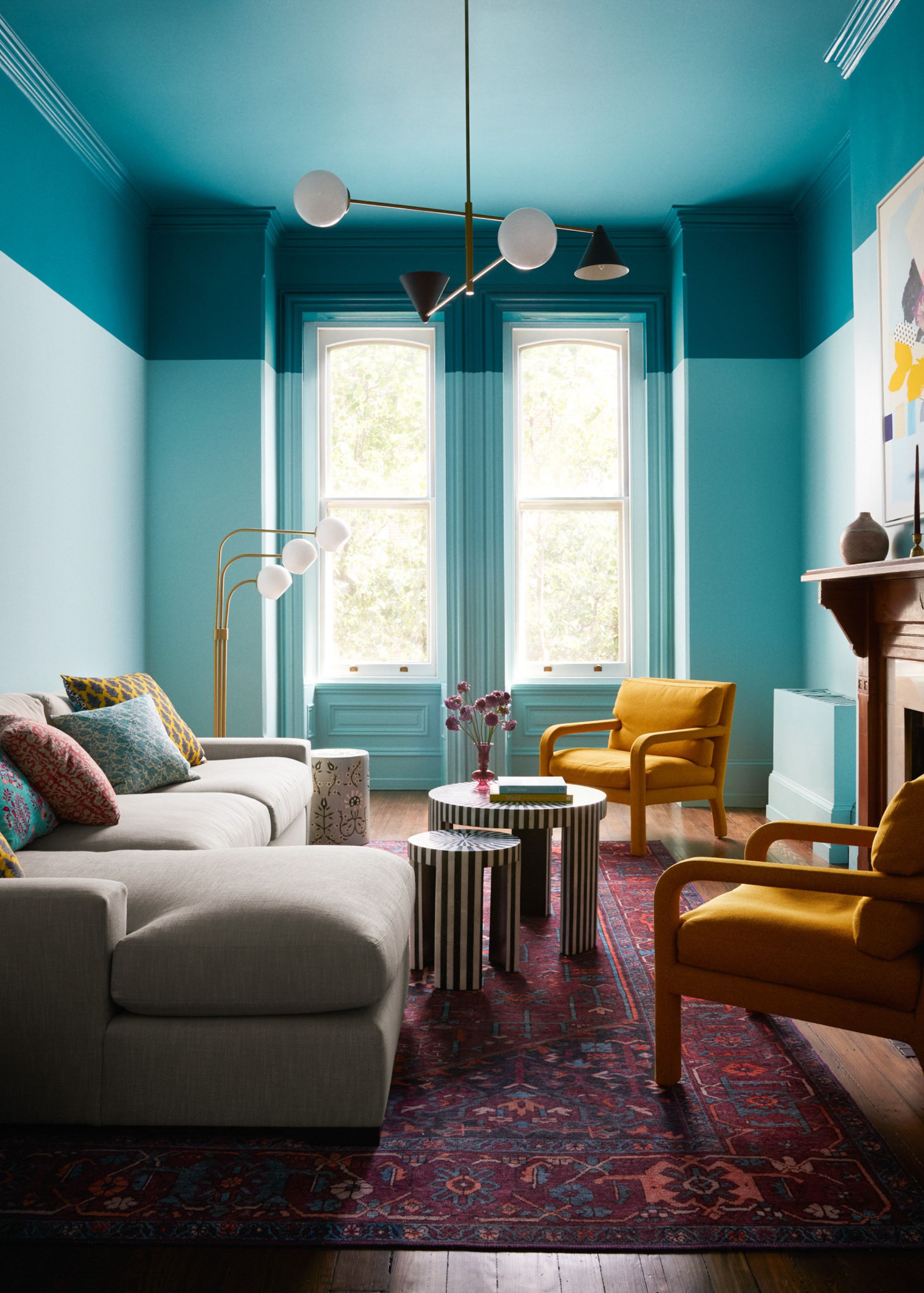 A modern living room with two-tone teal paints on the walls and ceiling. A red patterned rug and a cream chaise sofa with two mustard accent chairs.