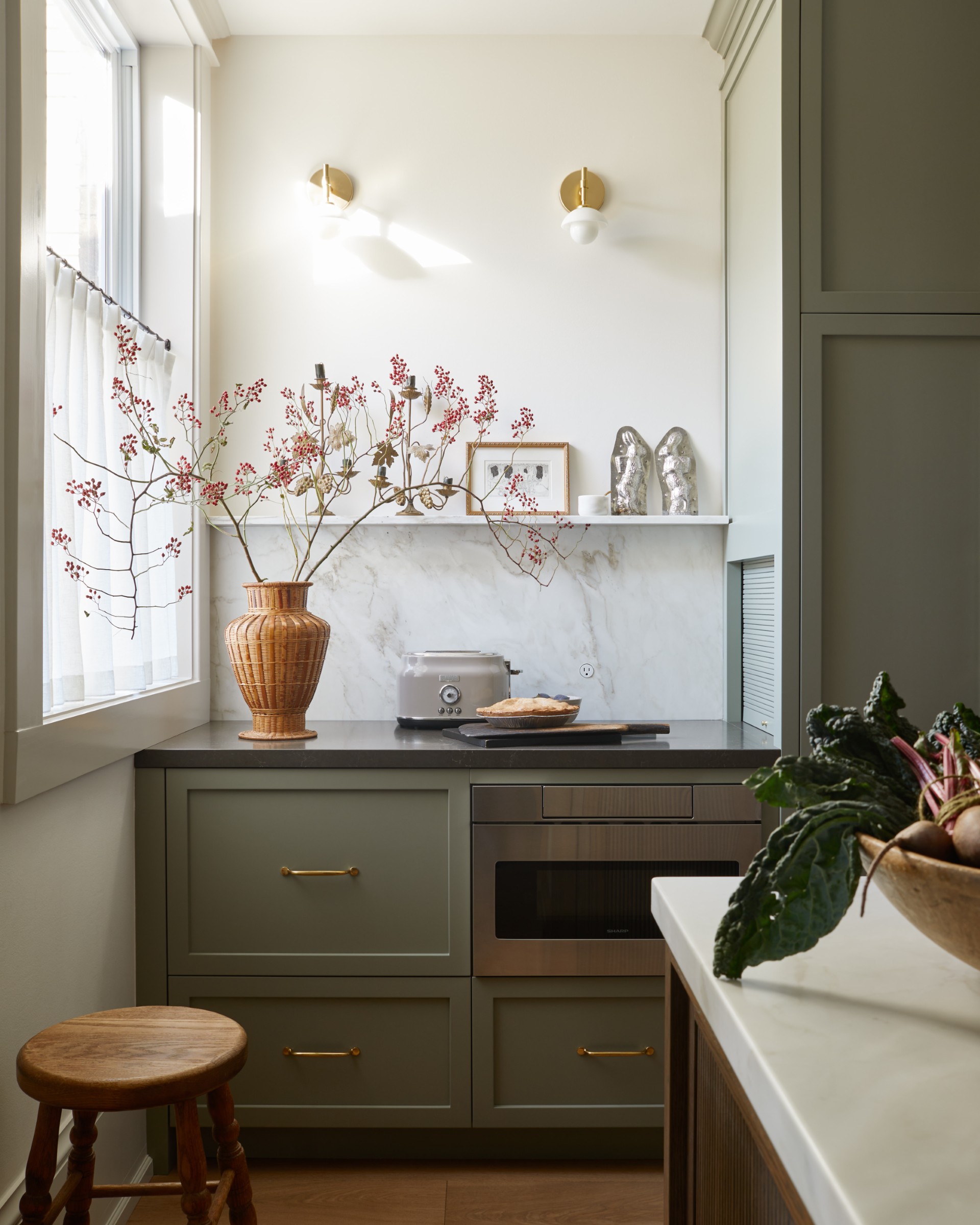 Sage green kitchen cabinets with white marble backsplash and white walls. Larger plant in wicker vase and grey toaster on couner. Next to window and two gold light fixtures on wall