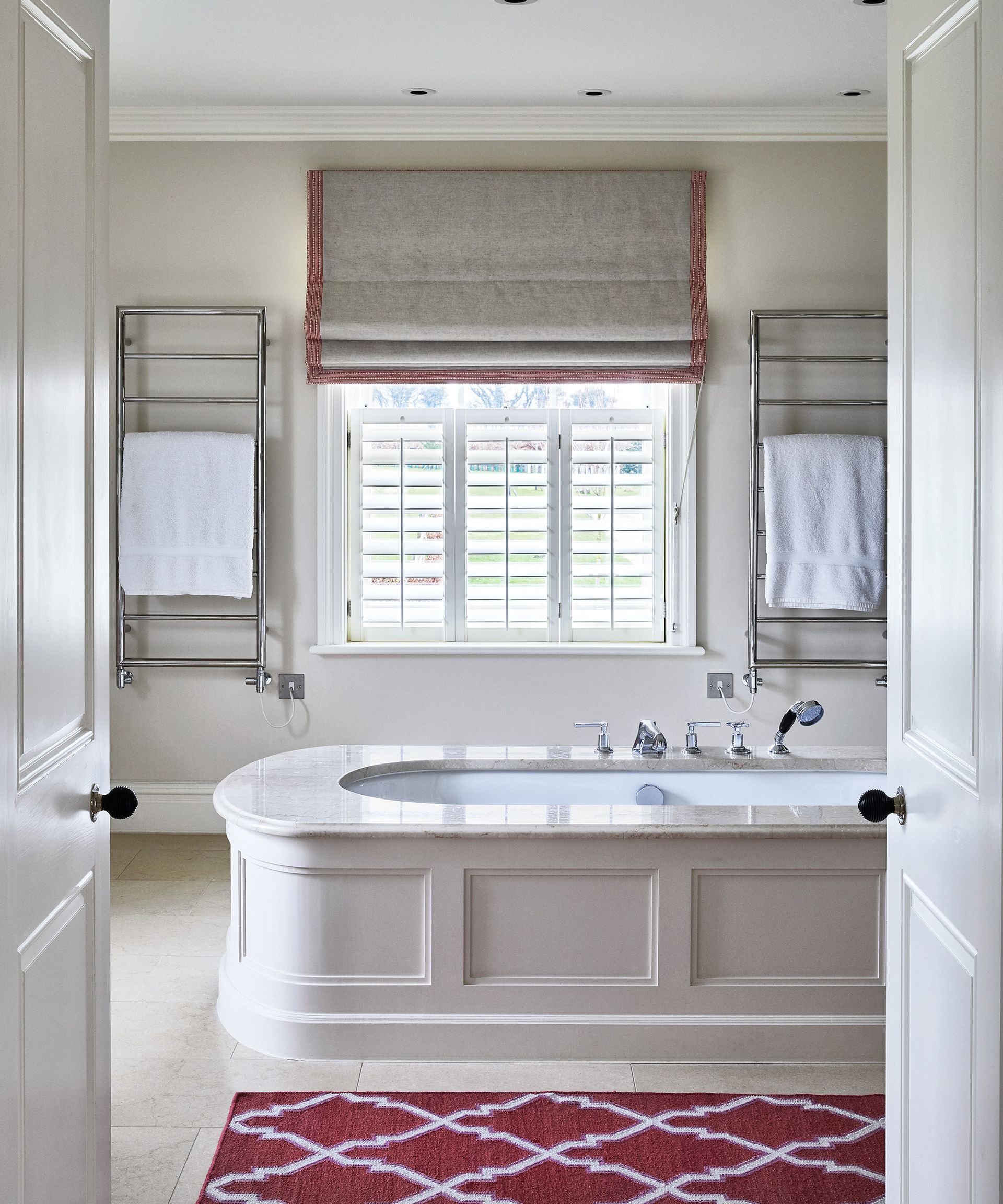 An example of bathroom color ideas showing a cream bathroom with a bath in the center next to a red and white rug