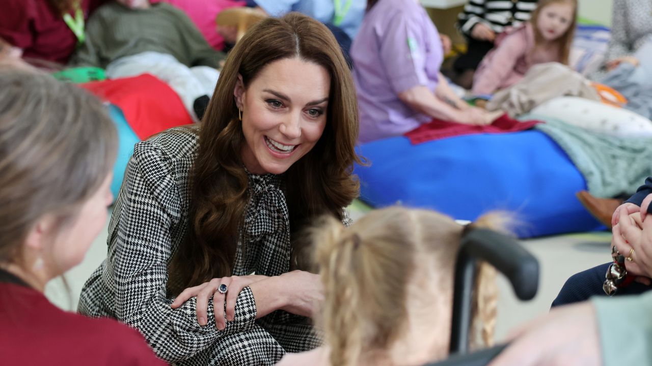 Kate Middleton wearing a black plaid dress with a tie neck kneeling on the floor talking to a little girl with families sitting behind her on the floor at a hospice