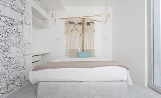 Bedroom with bed and white linen. There is a built-in dresses with drawers on the one side, a tapestry on the wall behind the bed and a aluminium door leading outside.
