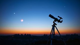 An image of a telescope standing in front of a sunset, with bright planets visible in the dimming sky
