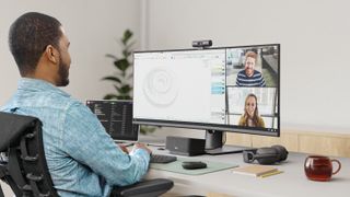 Man sitting at a desk with a computer screen on it, with Logitech webcam on top of the screen, and Logitech headphones on the desk