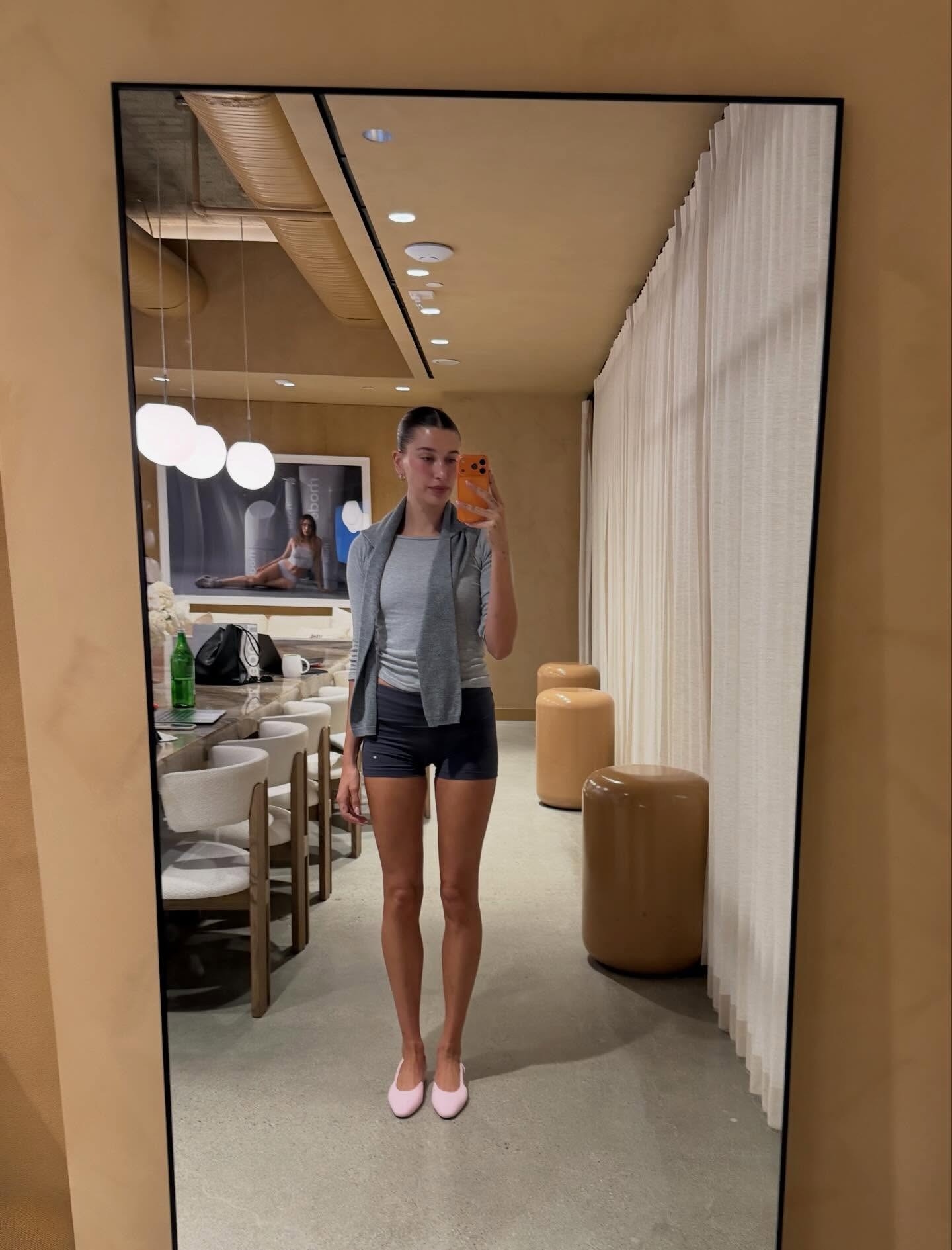 Hailey Bieber wears a gray shirt, gray shorts, and baby pink ballet flats.