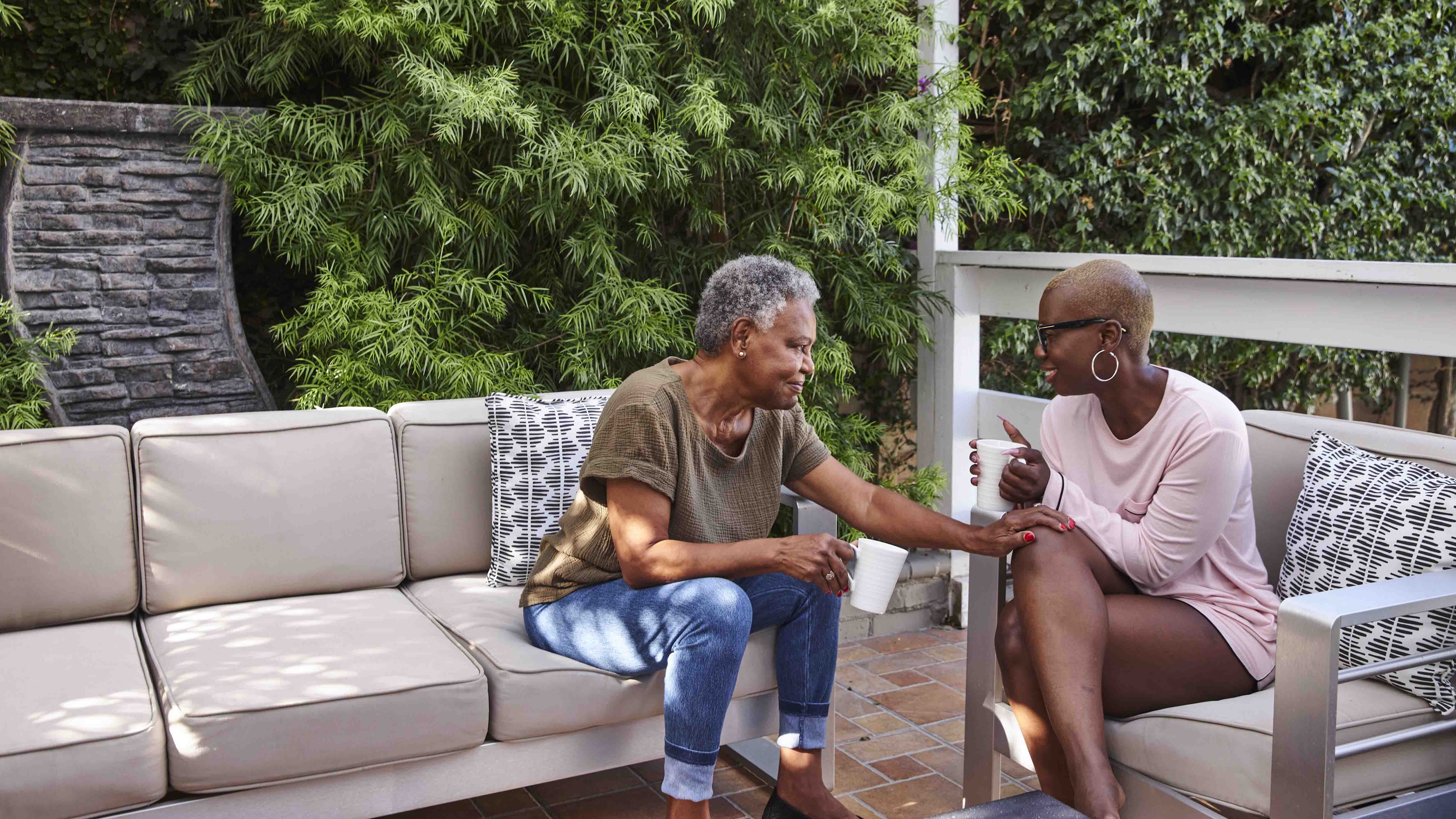 Two older women sitting outside on a deck and chat while drinking coffee.