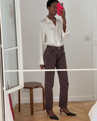 Paris-based fashion influencer Sylve Mus poses for a mirror selfie wearing a white satin button-down shirt, brown cuffed jeans, and black slingback heels.