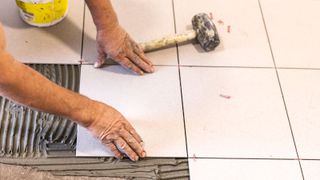 Hands laying square cream tiles onto grey tile adhesive with rubber mallet laying on tiles