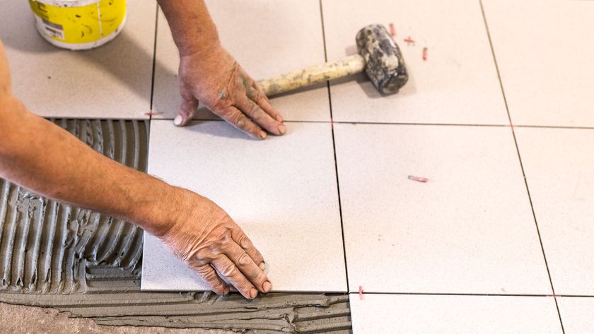 Hands laying square cream tiles onto grey tile adhesive with rubber mallet laying on tiles